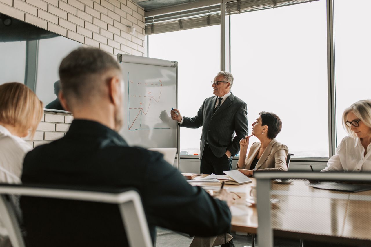 who-we-are Senior leader presenting growth charts in a business meeting with colleagues in a modern office setting.